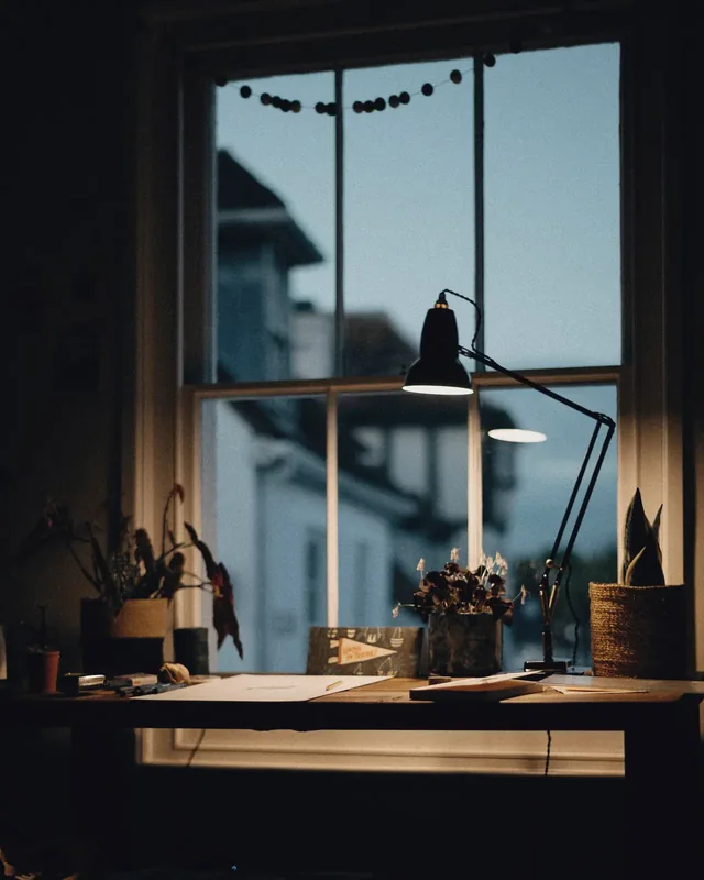 A desk by the window at dusk, the lamp switched on.